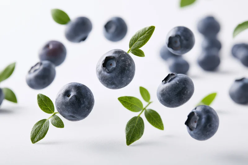 Blueberries floating midair with green leaves scattered on white surface