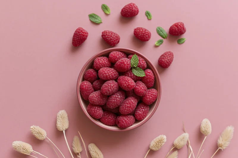 Fresh raspberries in a pink bowl with mint leaves and dried grasses on a pastel pink surface