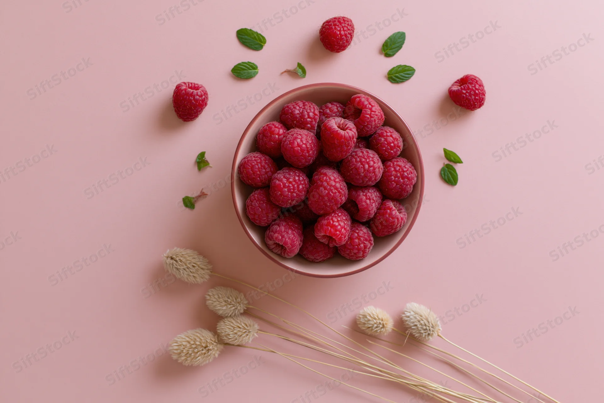 Fresh raspberries in a small bowl with mint leaves and dried flowers on a pink surface