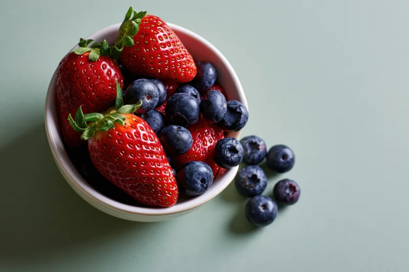 Strawberries and blueberries spilling from a white bowl on a mint green surface