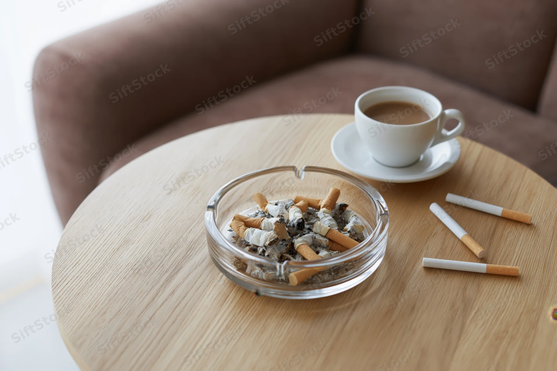 Glass ashtray filled with cigarette butts and coffee cup on wooden table near sofa