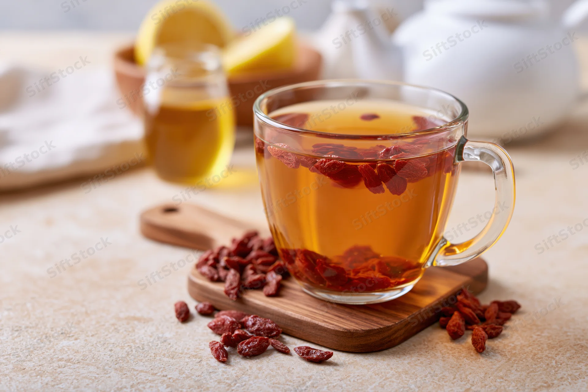 Glass mug of amber goji berry tea with dried berries floating, on wooden coaster with scattered berries, lemon and honey nearby