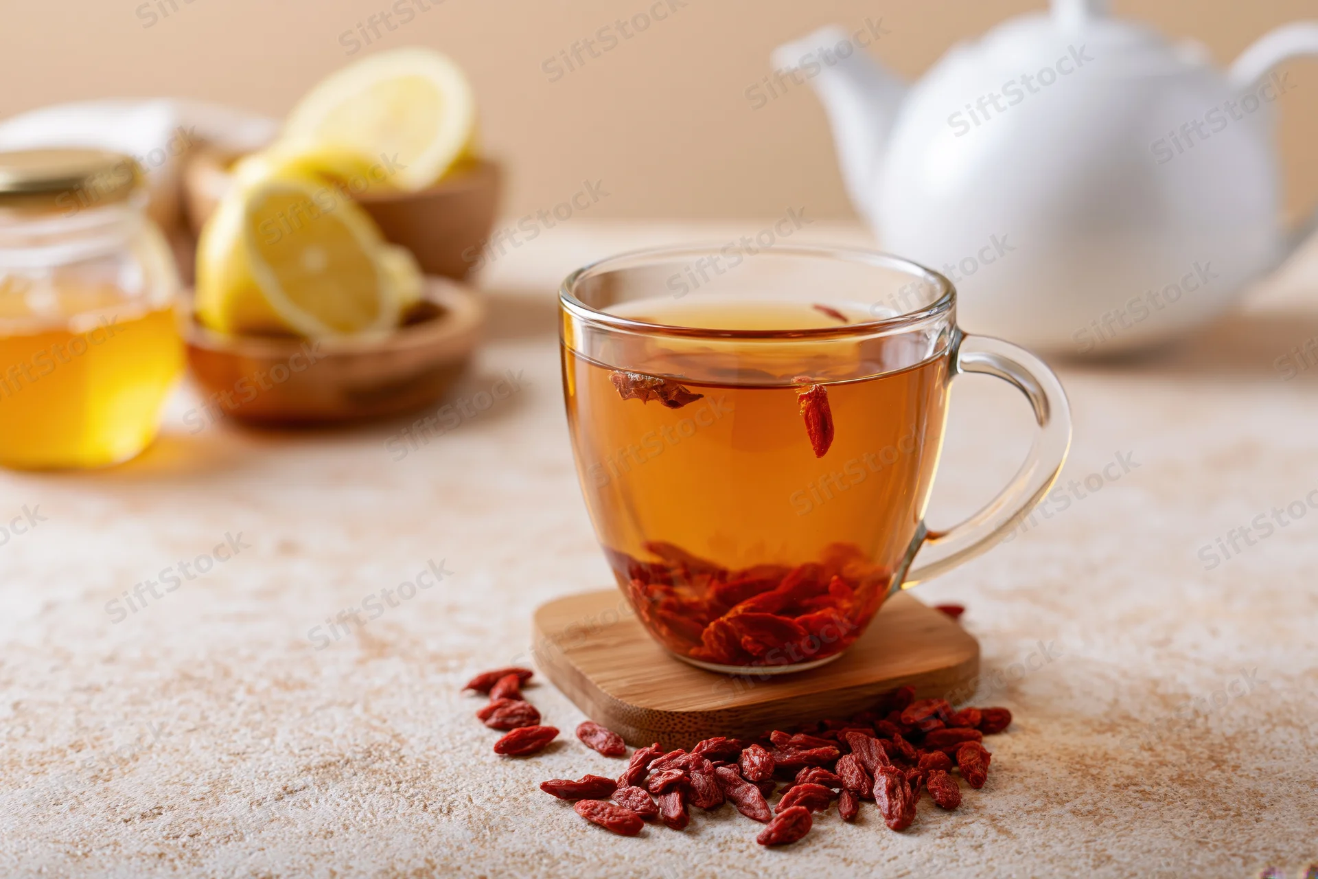 Glass mug of amber goji berry tea with dried berries floating, on a bamboo coaster beside honey and lemon slices