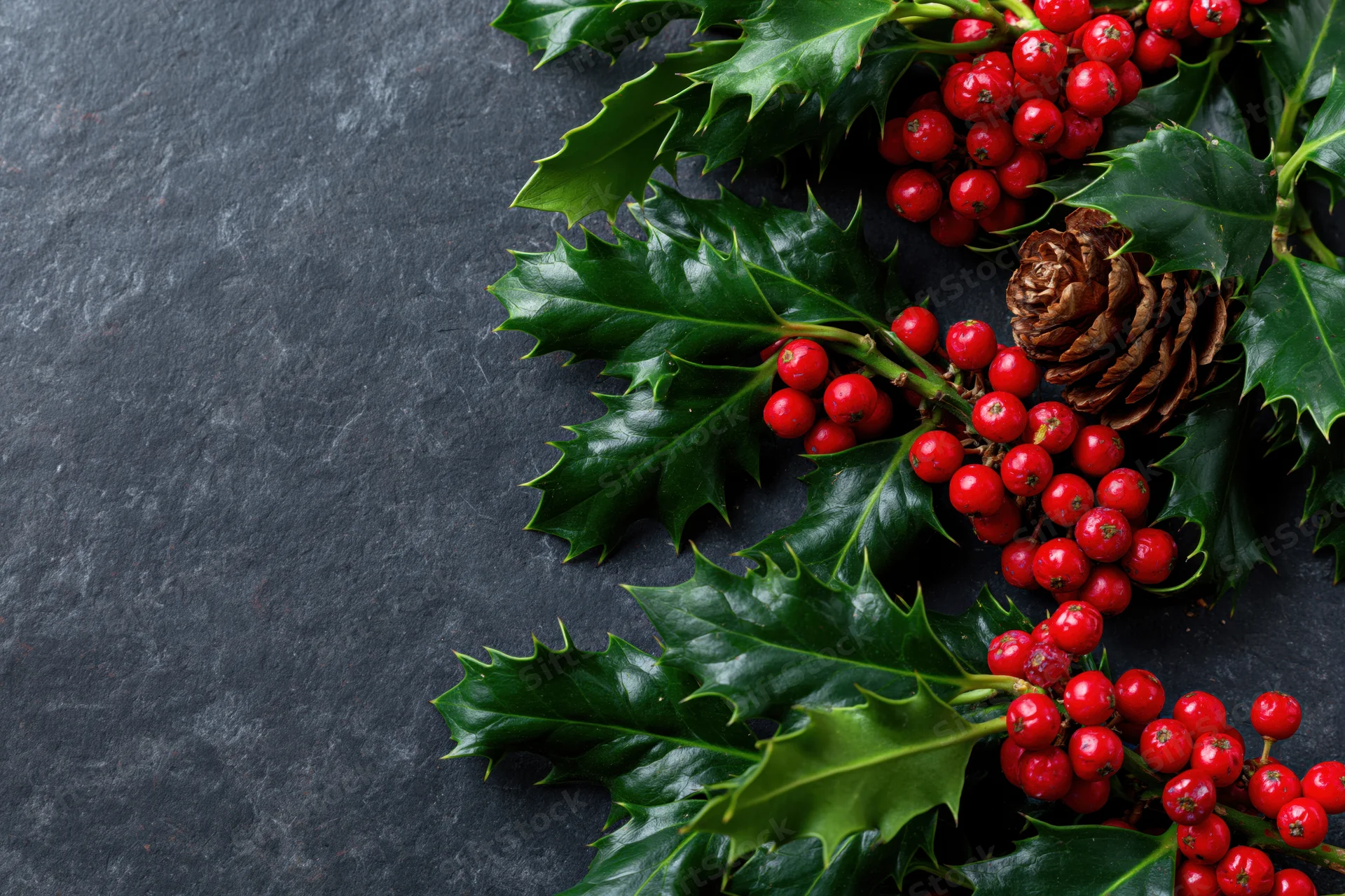 Holly branches with red berries and a pinecone arranged on a dark stone surface