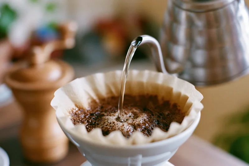 Hot water pouring from a gooseneck kettle into a paper filter filled with coffee grounds over a ceramic dripper