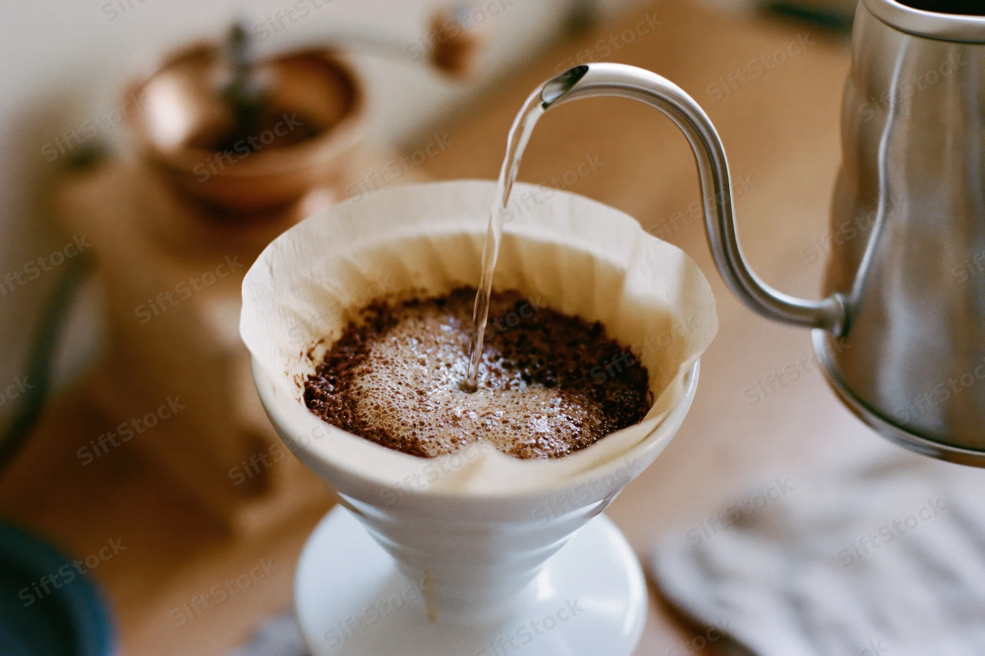 Hot water pouring from a gooseneck kettle into a paper filter over coffee grounds in a white ceramic funnel on a wooden countertop