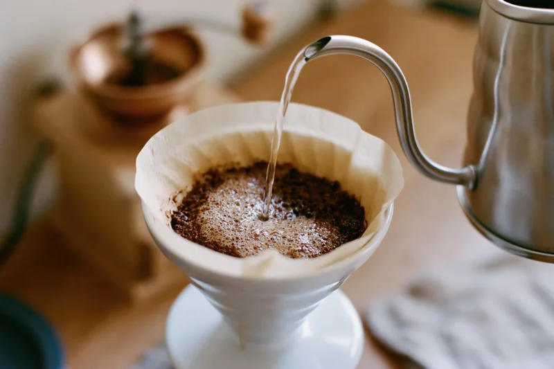Hot water pouring from a gooseneck kettle into a paper filter over coffee grounds in a white ceramic funnel on a wooden countertop