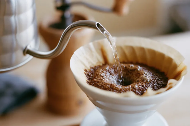 Hot water pouring from a gooseneck kettle into a paper filter filled with coffee grounds on a wooden surface