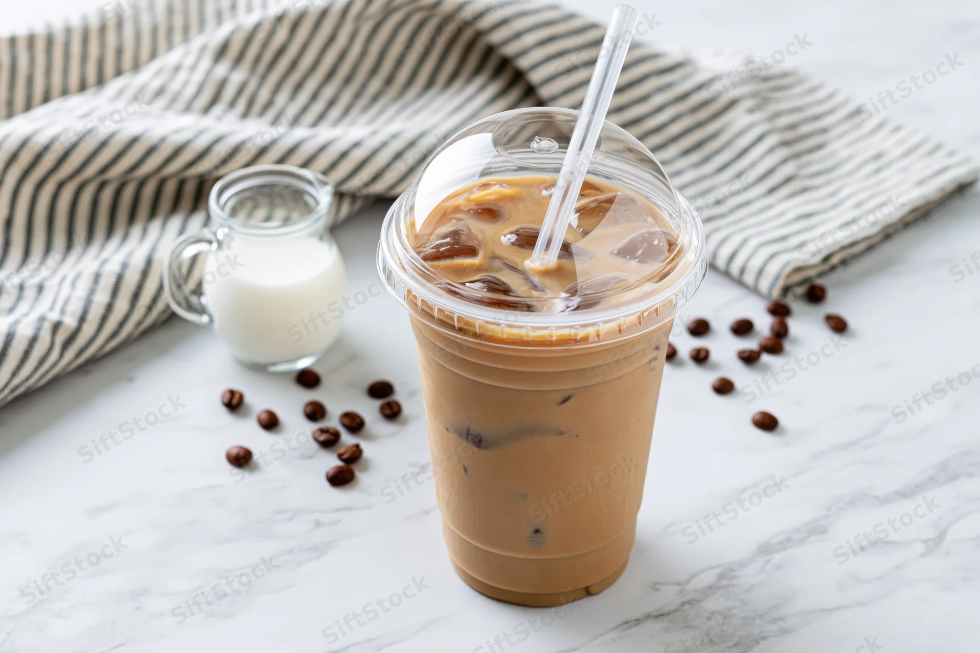 Iced coffee in a clear plastic takeaway cup with domed lid and straw on a marble surface beside a small glass jug of milk and scattered coffee beans