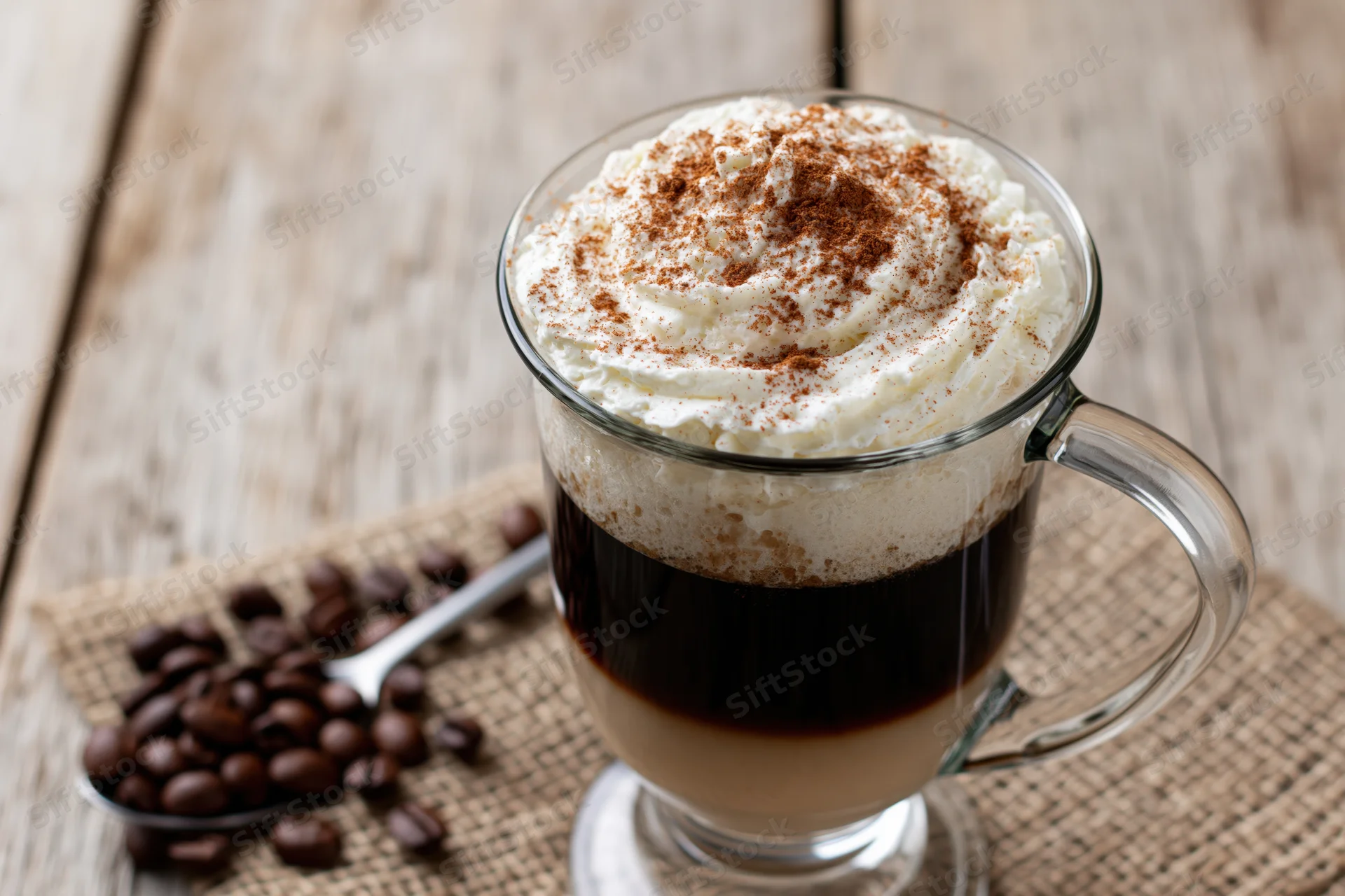 Irish coffee in a tall glass mug with whipped cream and cinnamon topping on a wooden table beside coffee beans and a spoon