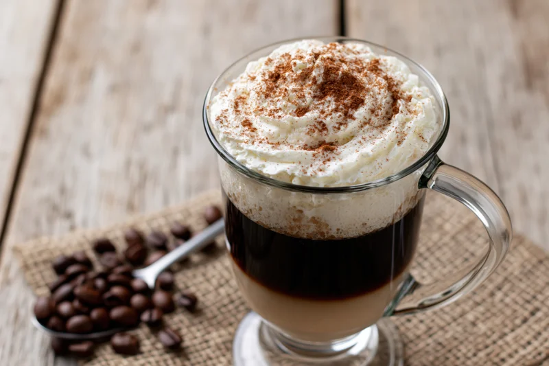 Irish coffee in a tall glass mug with whipped cream and cinnamon topping on a wooden table beside coffee beans and a spoon