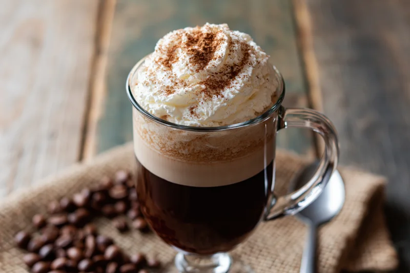 Tall glass mug of layered irish coffee with whipped cream and cinnamon on a wooden table with coffee beans and a spoon