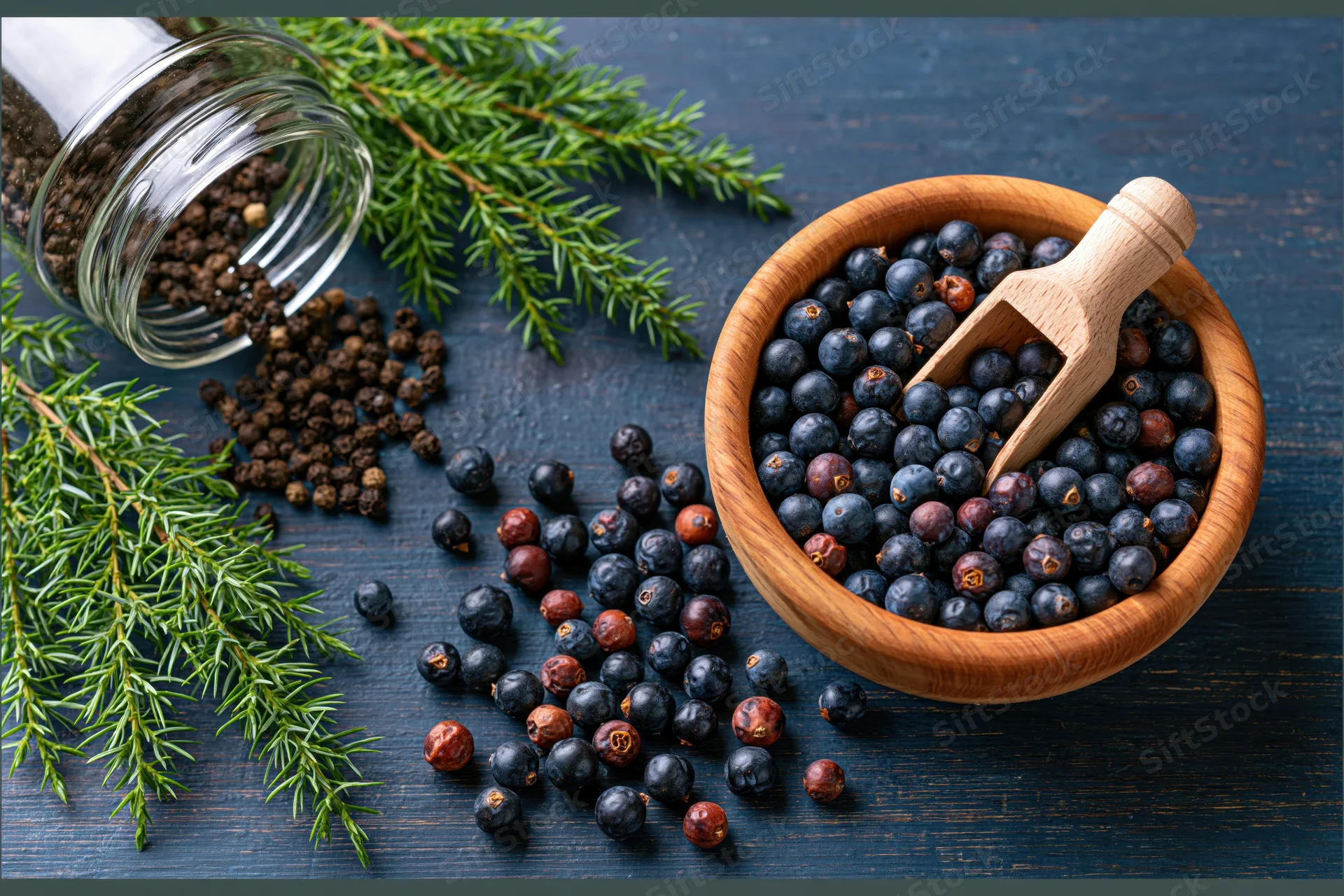 Juniper berries in a wooden bowl with a small wooden scoop on a dark wooden surface surrounded by evergreen branches and scattered peppercorns