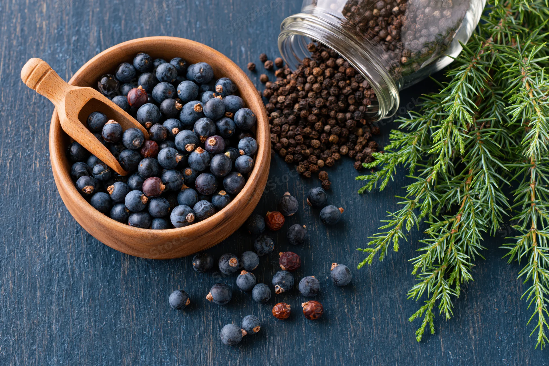 Juniper berries in a wooden bowl with a small wooden scoop beside a glass jar and pine sprig on dark wood surface