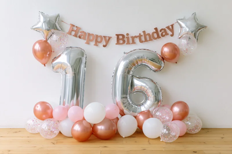 Silver foil number balloons spelling 16 arranged against a white wall with pink and rose gold balloons and a happy birthday banner on a wooden floor