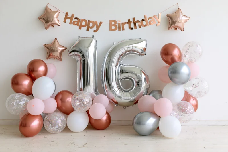 Silver foil number balloons spelling 16 surrounded by rose gold pink and white balloons with a happy birthday banner above on a light wooden floor against a white wall