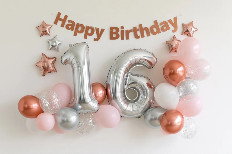Silver foil number balloons spelling 16 with pink and rose gold balloons and a happy birthday banner on a white wall