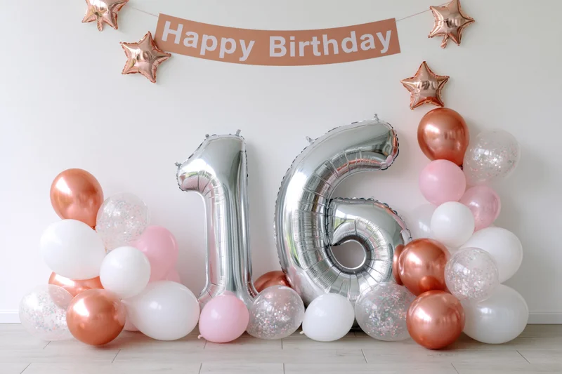 Silver foil number balloons spelling 16 arranged against a white wall with rose gold and pink balloons and a happy birthday banner above