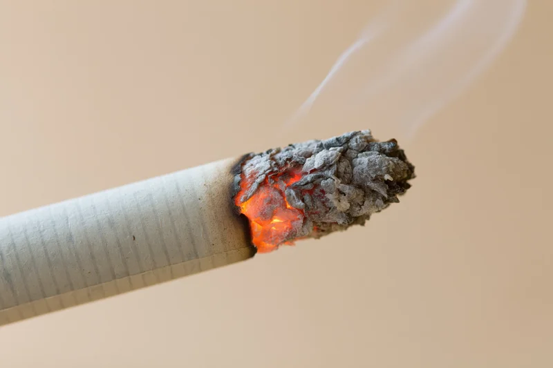 Closeup of lit cigarette tip with glowing ember and rising smoke against plain background
