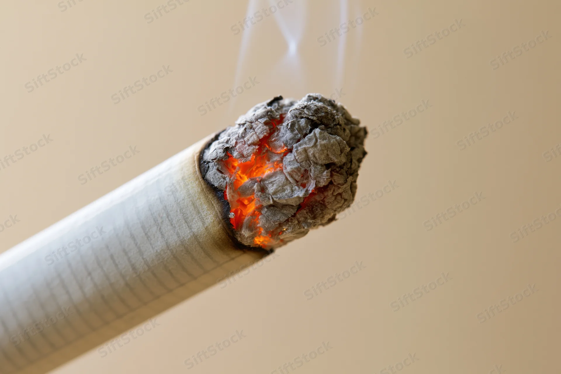 Lit cigarette tip glowing with orange ember and ash in closeup against beige background
