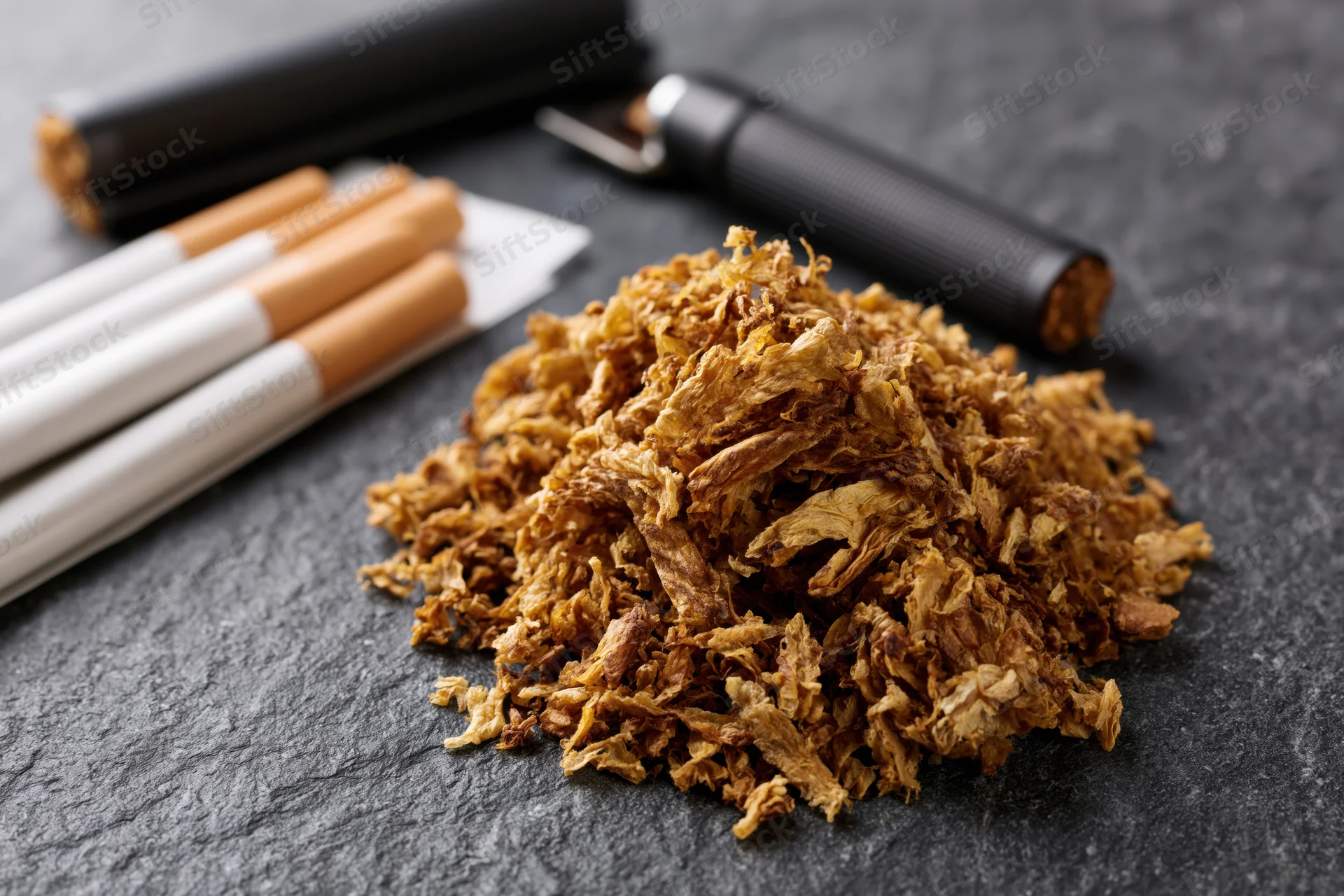 Pile of loose rolling tobacco on dark slate surface with cigarettes and cutting tool nearby