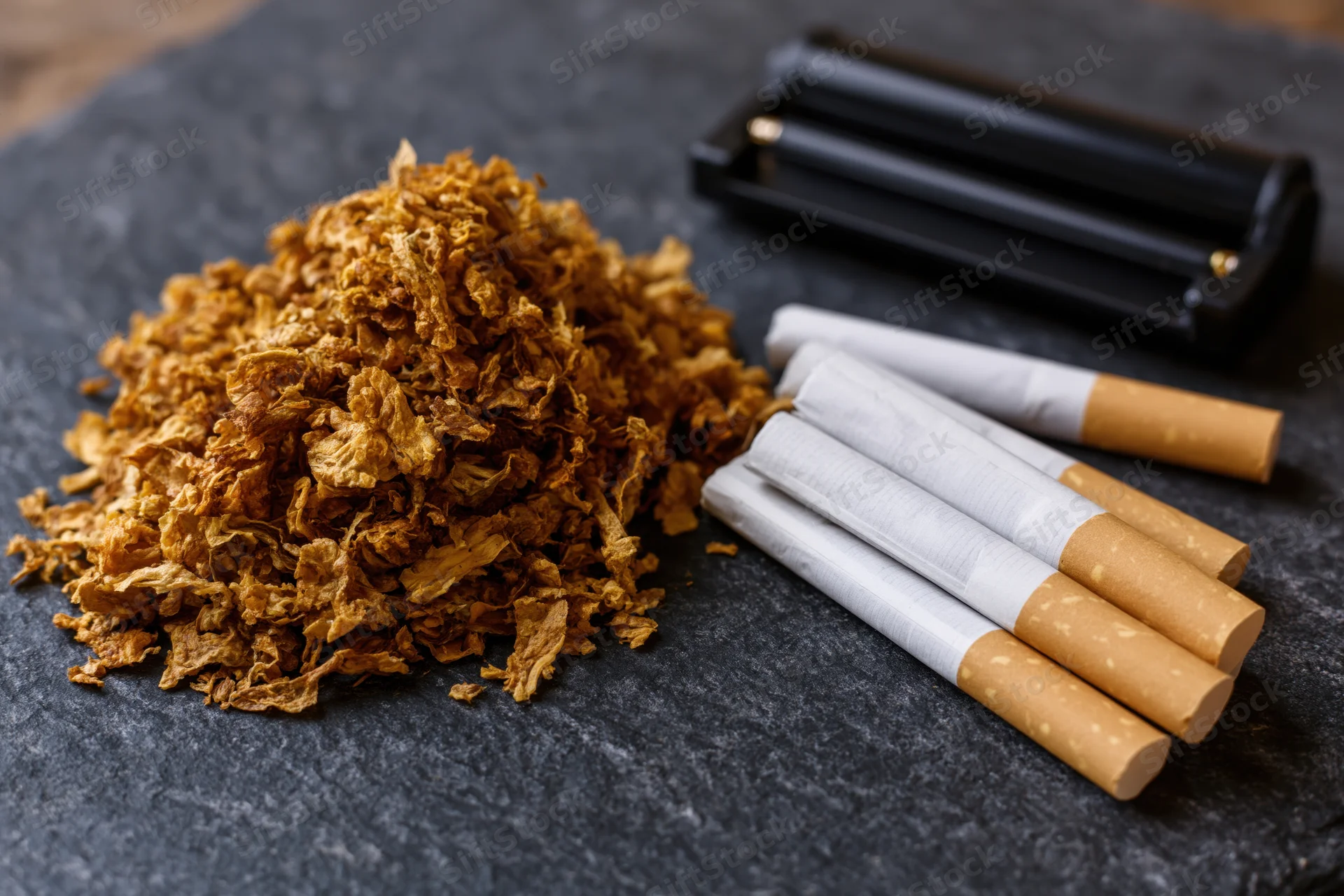Loose tobacco piled next to rolled cigarettes and a rolling machine on dark slate surface