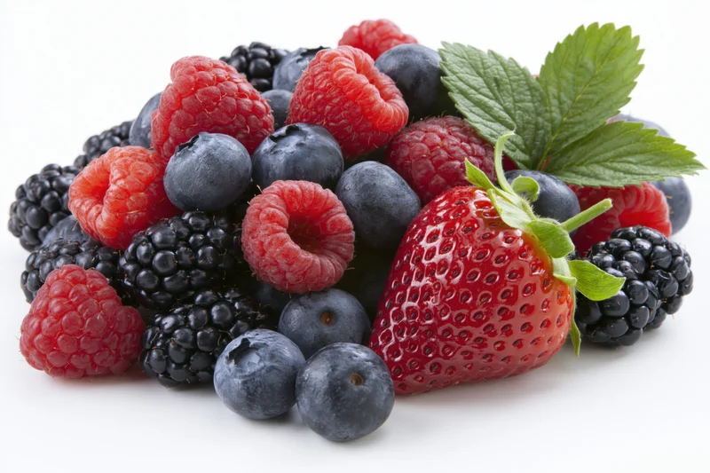 A pile of fresh strawberries, raspberries, blueberries and blackberries with a green leaf on a white surface