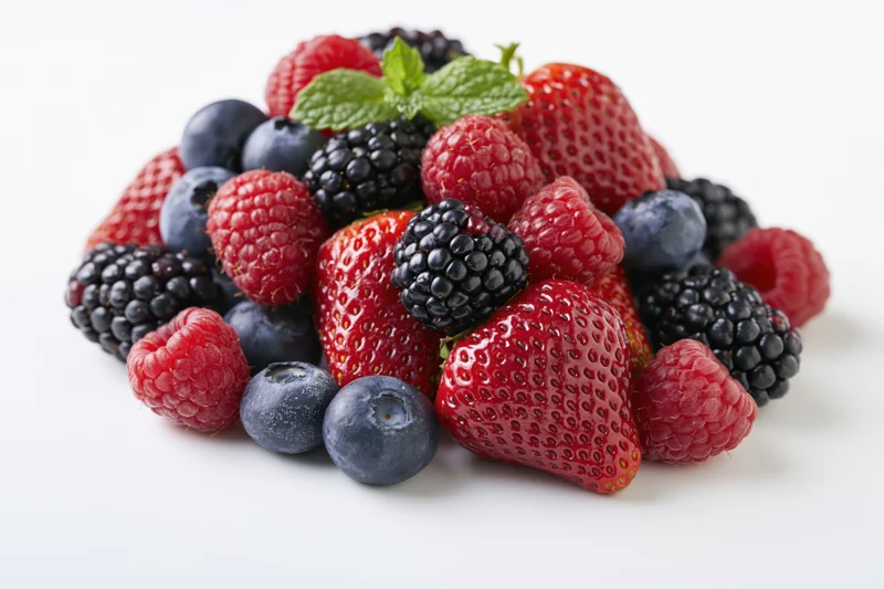 A pile of mixed fresh berries including strawberries, raspberries, blueberries and blackberries with a mint leaf on a white background