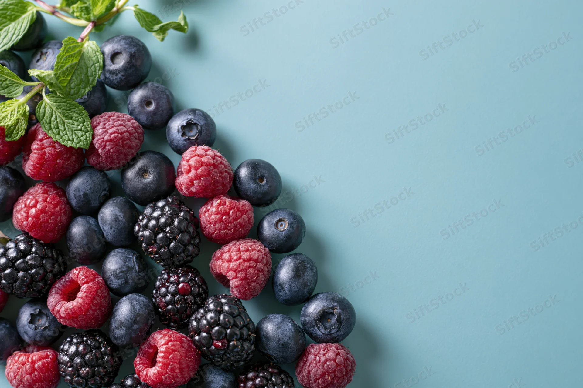 Mixed fresh berries including blueberries raspberries and blackberries arranged with mint leaves on a teal surface