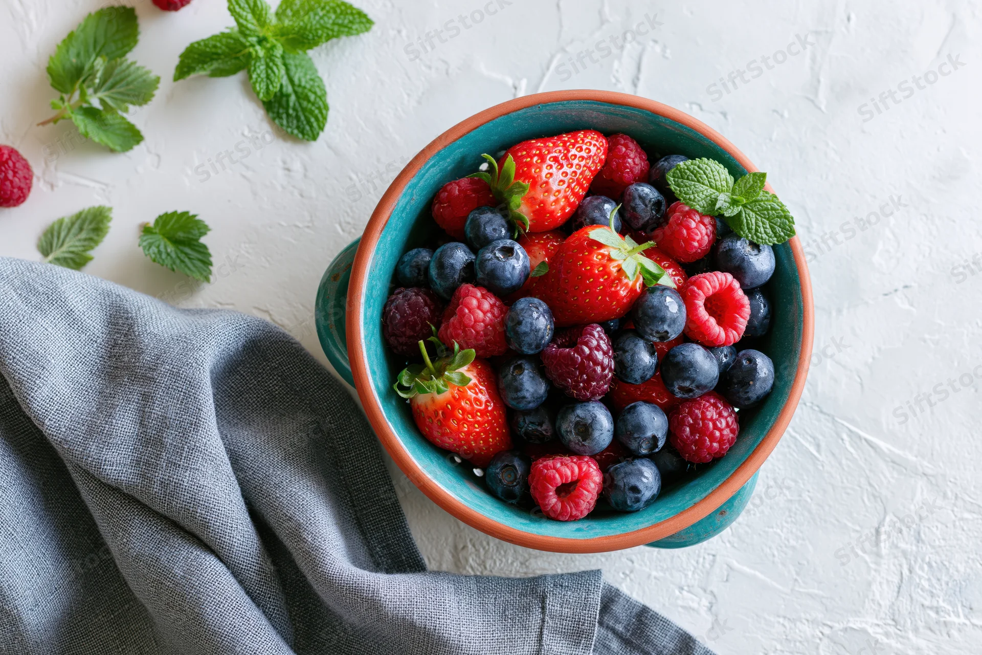 Mixed fresh berries in a ceramic bowl with mint on a white surface