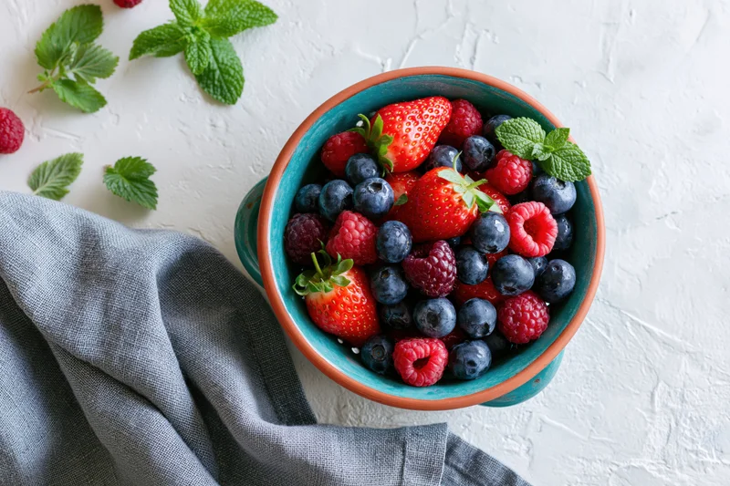 Mixed fresh berries in a ceramic bowl with mint on a white surface