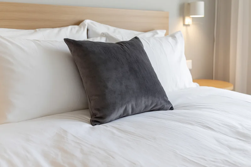 Neatly made hotel bed with white bedding and a dark gray decorative pillow against a wooden headboard