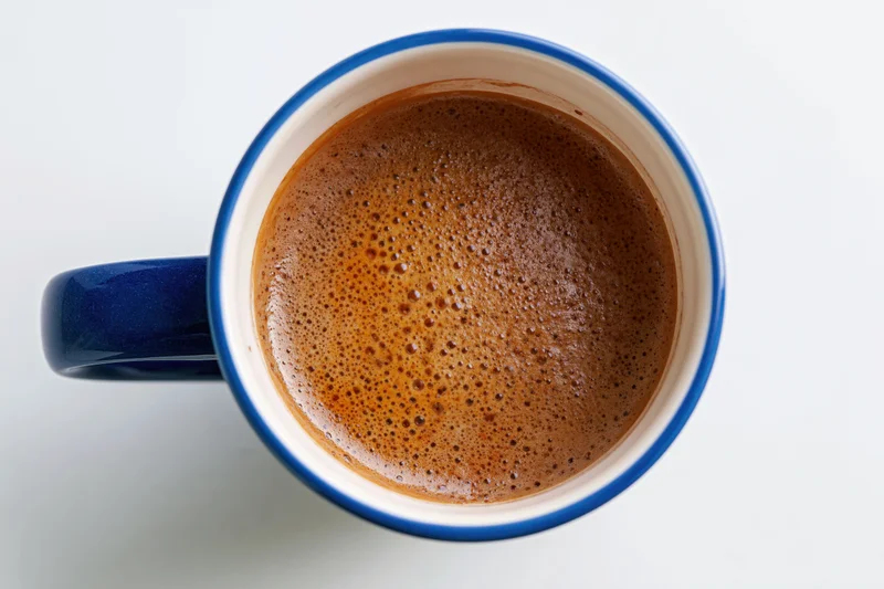 Overhead closeup of a blue ceramic coffee mug filled with frothy coffee on a white surface