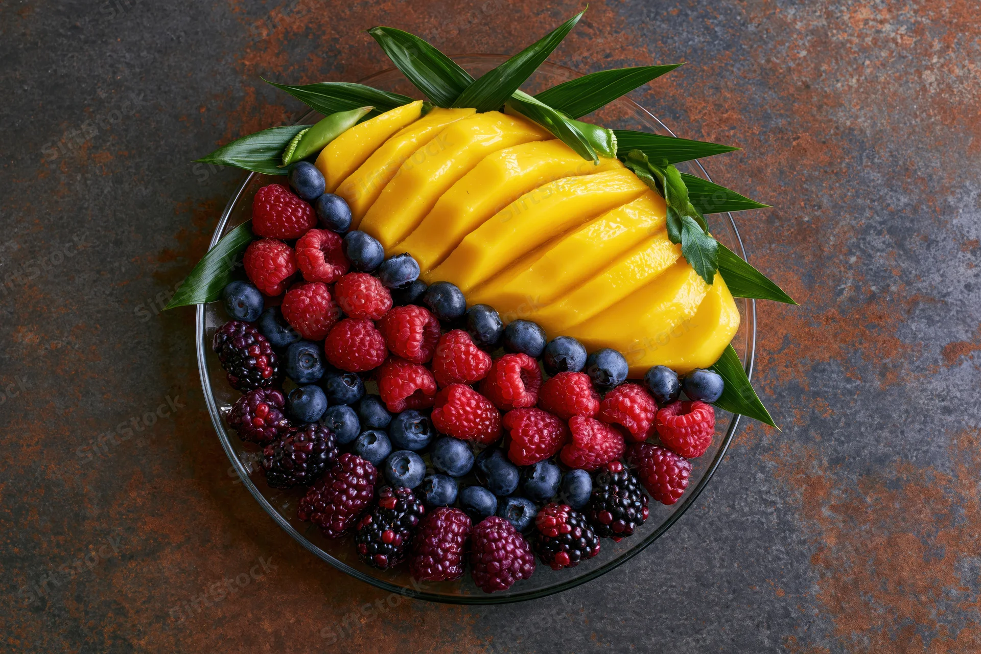 Overhead flat-lay of a glass plate with sliced mango and mixed berries arranged with green leaves on a dark rustic surface
