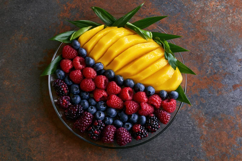 Overhead flat-lay of a glass plate with sliced mango and mixed berries arranged with green leaves on a dark rustic surface