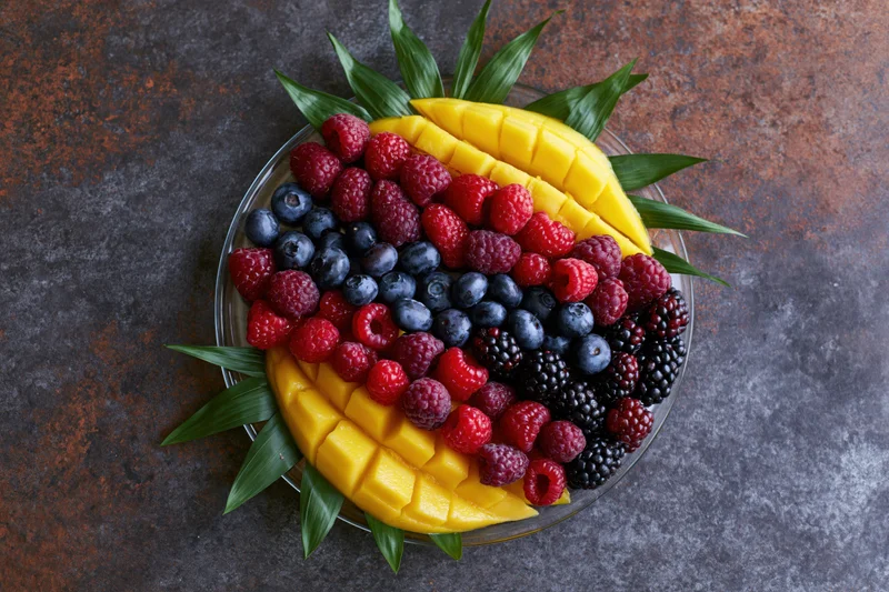 Overhead flat-lay of a glass plate with sliced mango and mixed berries arranged with green leaves on a dark textured surface