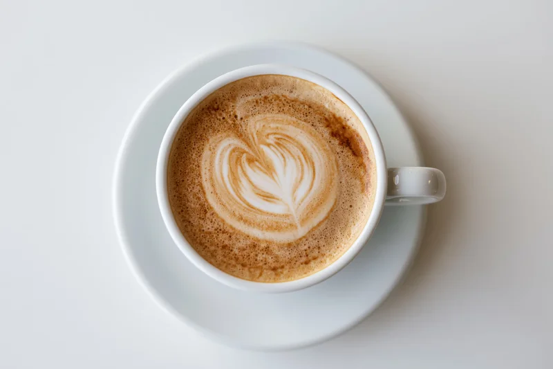 Overhead view of a white ceramic cappuccino cup with heart-shaped latte art on a saucer against a white background