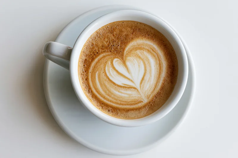 Overhead view of a white ceramic cappuccino cup with heart-shaped latte art on a saucer against a plain white background