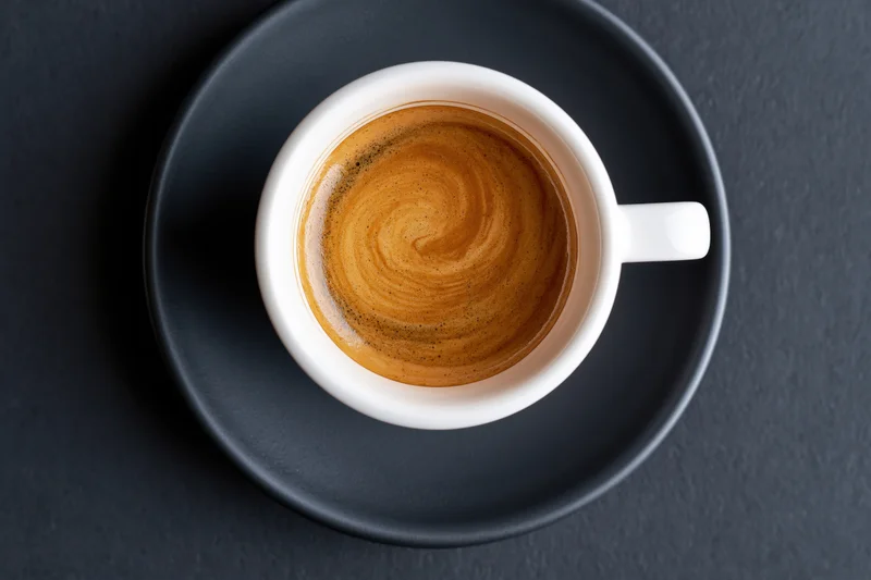 Overhead view of a white ceramic espresso cup with crema swirl on a dark saucer