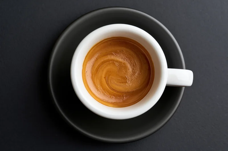 Overhead view of a white ceramic espresso cup with crema swirl on a black saucer and dark surface