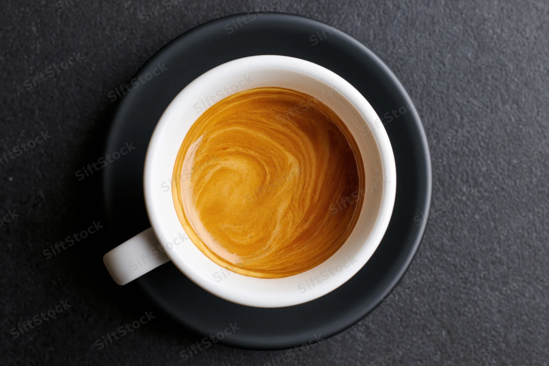 Overhead view of a white ceramic espresso cup with latte art on a black saucer on dark stone surface