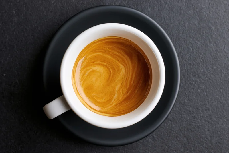 Overhead view of a white ceramic espresso cup with latte art on a black saucer on dark stone surface