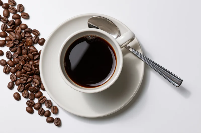 Overhead view of a white ceramic coffee cup filled with black coffee on a saucer with a spoon and scattered coffee beans on a white surface