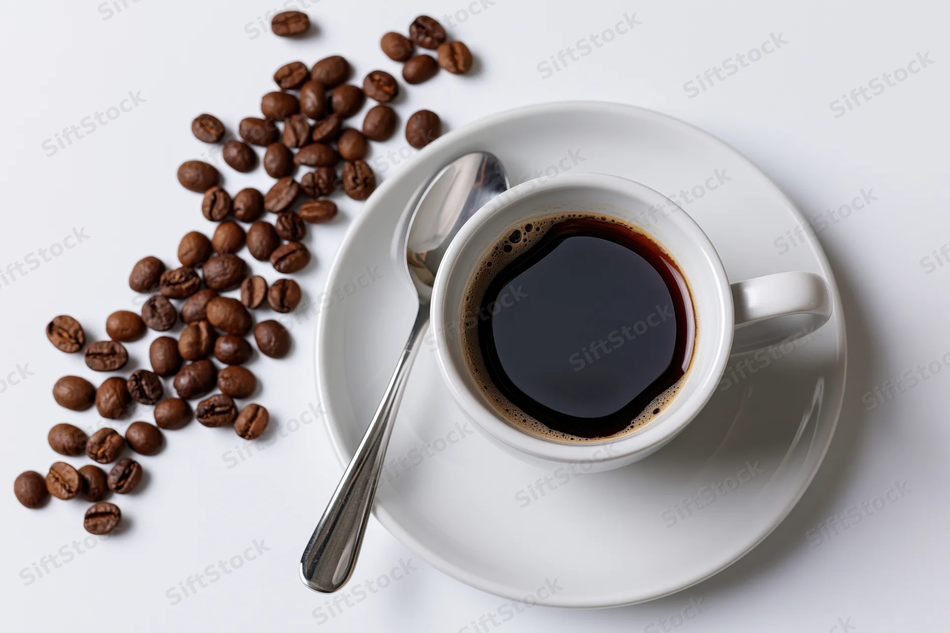 White ceramic coffee cup filled with black coffee on saucer with spoon and scattered beans on white surface
