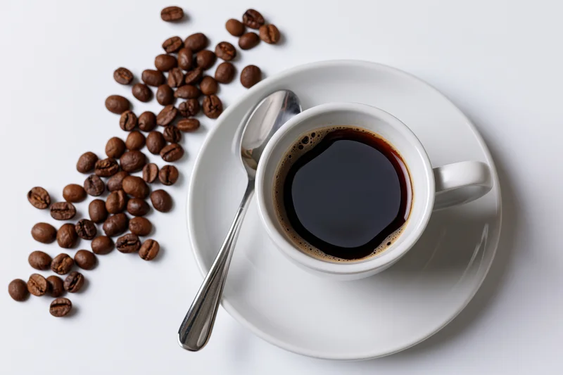 White ceramic coffee cup filled with black coffee on saucer with spoon and scattered beans on white surface