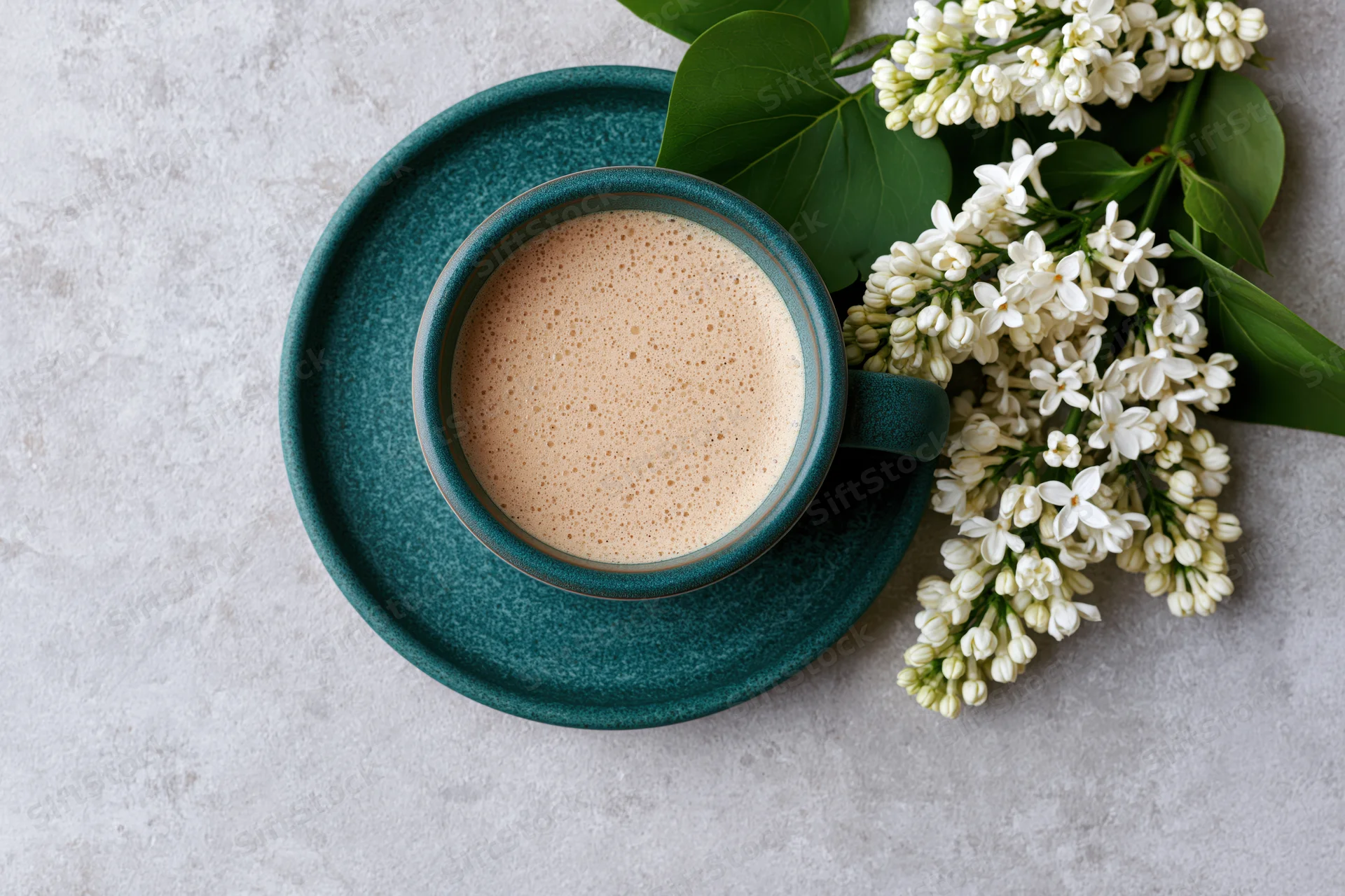 Teal ceramic coffee cup with frothy beverage and white lilac flowers on gray surface in flat lay arrangement