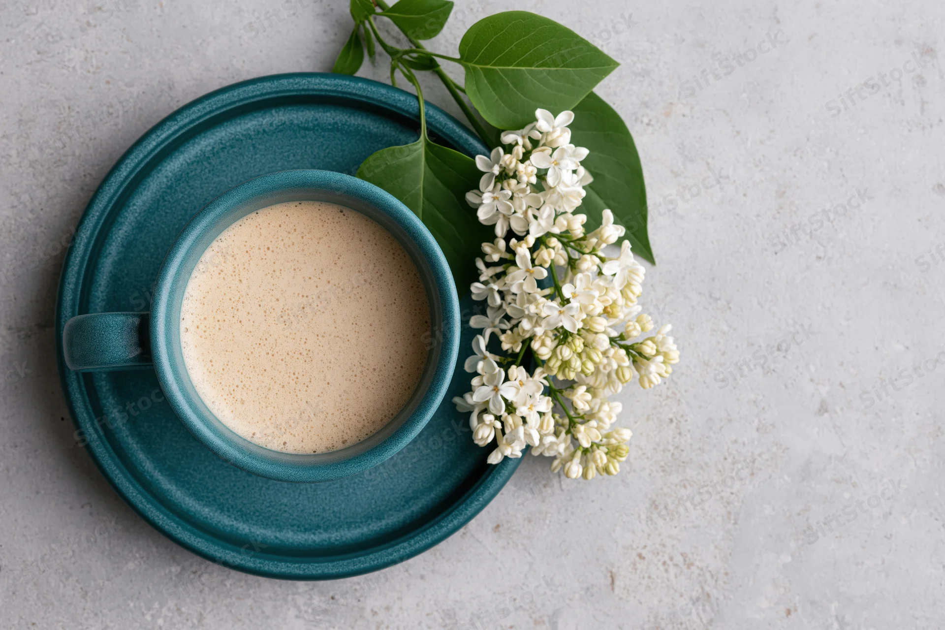 Teal ceramic coffee cup with frothed milk and white lilac flowers on a gray stone surface