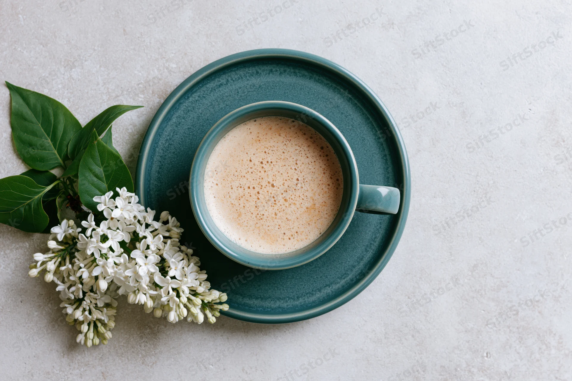 Overhead flat lay of a teal ceramic coffee cup with frothy latte on a saucer beside white lilac flowers on a light textured surface