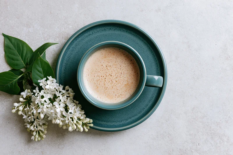 Overhead flat lay of a teal ceramic coffee cup with frothy latte on a saucer beside white lilac flowers on a light textured surface