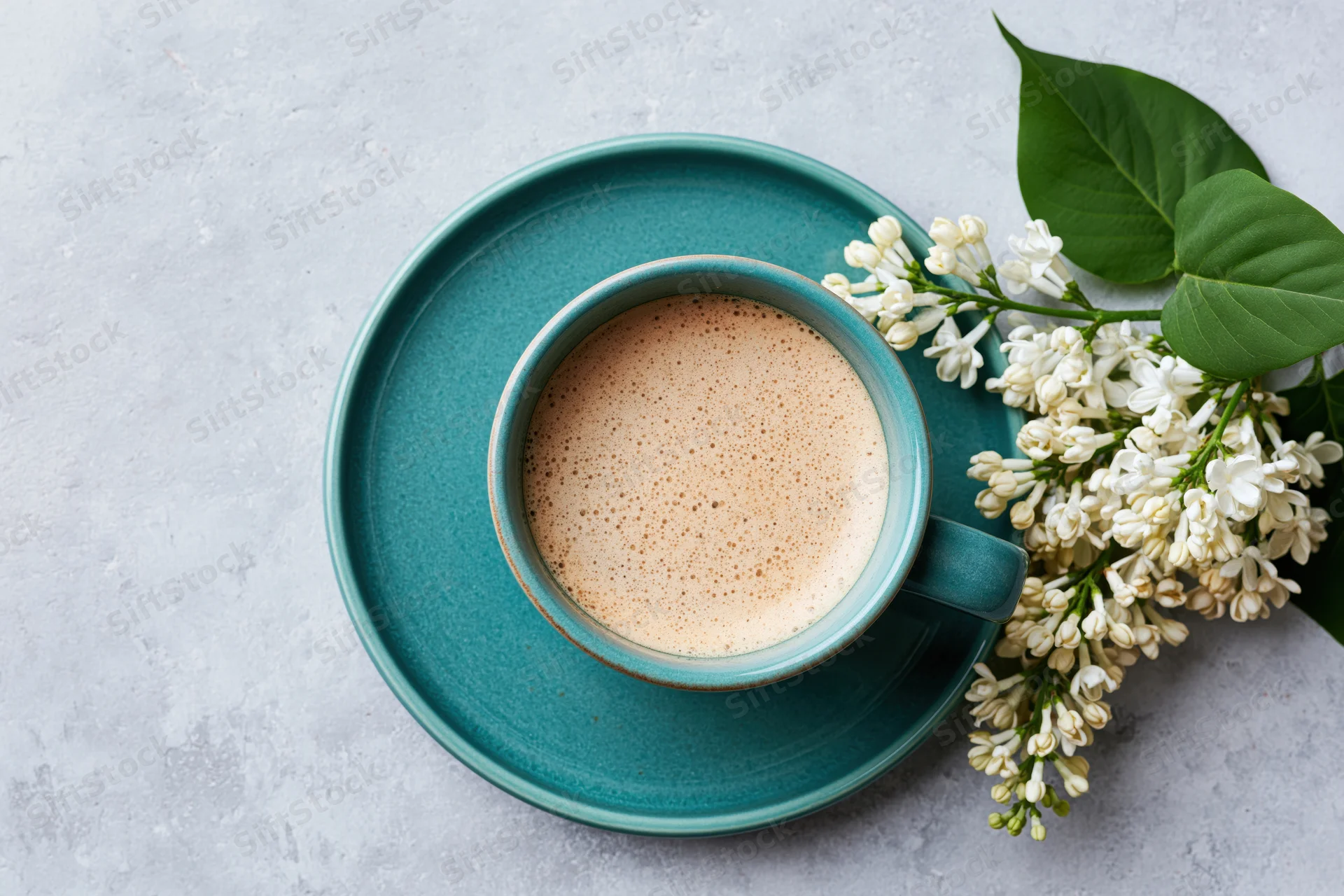 Teal ceramic coffee cup with frothy latte and white lilac flowers on gray surface